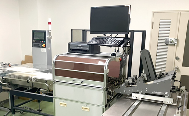 こちらはアウトソーシングのページとなります。これは検査装置の写真です。中央の装置で番号を読み、左の装置で重量を量ります。そしてそれらをPCで制御します。